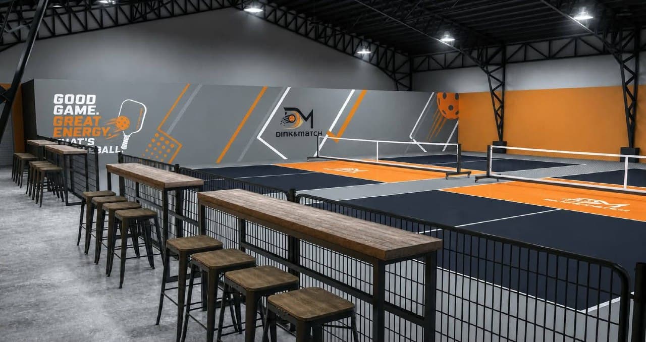 Indoor pickleball courts in navy and orange, with the Dink&Match mural along the far wall and bar seating beside the court edge
